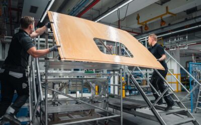 Zwei Monteure tragen ein Fahrerhausdach aus Holz auf den Kabinenrohbau hinauf