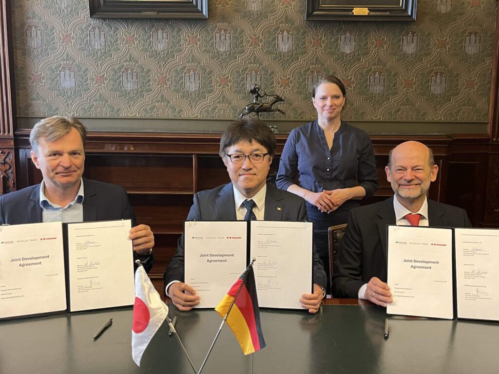 Volker Ebeling (MB Energy), Kei Nomura (Kawasaki Heavy Industries) und Dr. Manfred Schuckert (Daimler Truck), von links nach rechts sitzen, unterzeichnen im Beisein von Dr. Melanie Leonhard, Senatorin der Hamburger Behörde für Wirtschaft und Innovation (im Hintergrund stehend) eine Entwicklungsvereinbarung (Joint Development Agreement, JDA) zum Aufbau einer Lieferkette für Flüssigwasserstoff