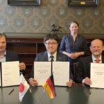 Volker Ebeling (MB Energy), Kei Nomura (Kawasaki Heavy Industries) und Dr. Manfred Schuckert (Daimler Truck), von links nach rechts sitzen, unterzeichnen im Beisein von Dr. Melanie Leonhard, Senatorin der Hamburger Behörde für Wirtschaft und Innovation (im Hintergrund stehend) eine Entwicklungsvereinbarung (Joint Development Agreement, JDA) zum Aufbau einer Lieferkette für Flüssigwasserstoff
