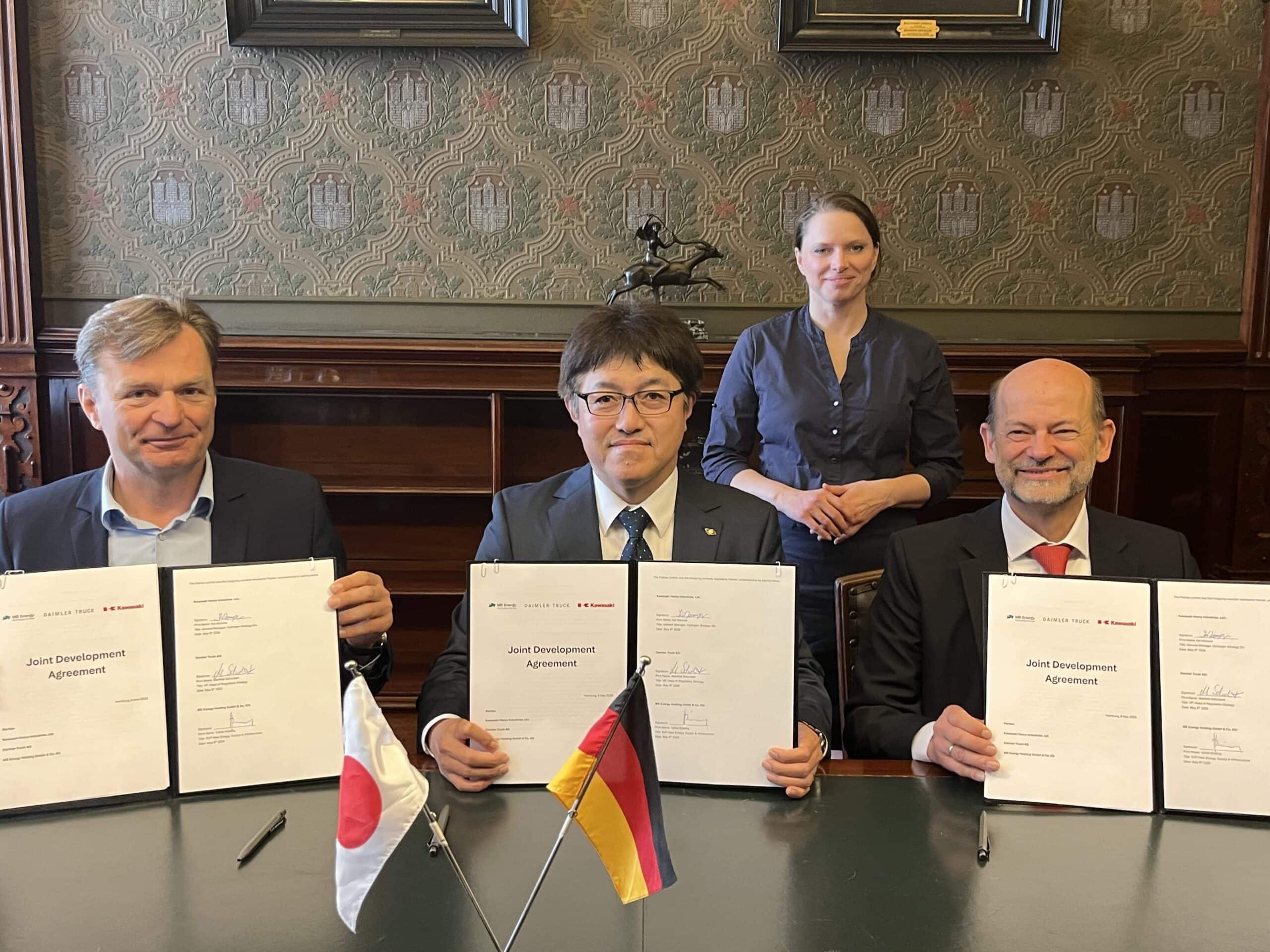 Volker Ebeling (MB Energy), Kei Nomura (Kawasaki Heavy Industries) und Dr. Manfred Schuckert (Daimler Truck), von links nach rechts sitzen, unterzeichnen im Beisein von Dr. Melanie Leonhard, Senatorin der Hamburger Behörde für Wirtschaft und Innovation (im Hintergrund stehend) eine Entwicklungsvereinbarung (Joint Development Agreement, JDA) zum Aufbau einer Lieferkette für Flüssigwasserstoff