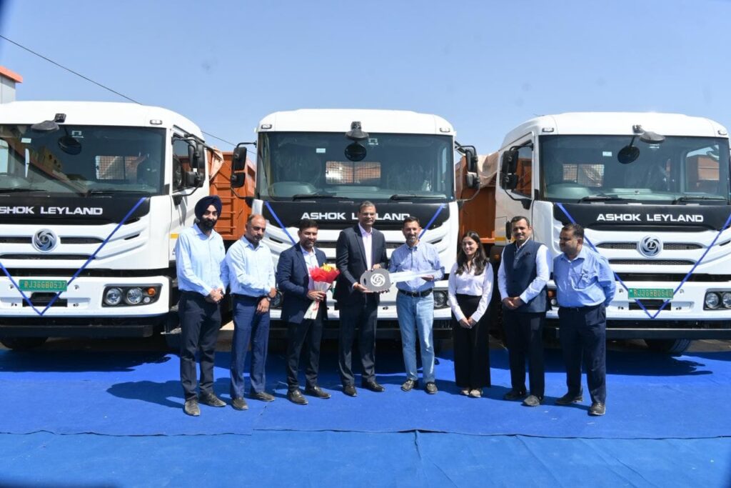 Eine Personengruppe mit Mitarbeitern von Ashok Leyland und ASAT Logistics vor drei schweren E-Lkw