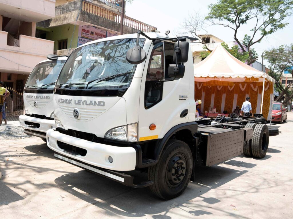 Zwei batterieelektrische Lkw vom Typ BOSS Electric von Ashok Leyland nebeneinander geparkt vor einem Gebäudekomplex; reine Fahrgestelle ohne Aufbau
