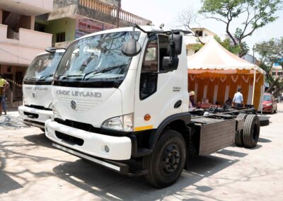 Zwei batterieelektrische Lkw vom Typ BOSS Electric von Ashok Leyland nebeneinander geparkt vor einem Gebäudekomplex; reine Fahrgestelle ohne Aufbau