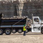 Schwerer Baustellen-Lkw der US-Marke Western Star mit überblendeter Illustration des eingebauten Motors der neuen Gen 6-Generation von Detroit Diesel; im Hintergrund eine Felswand, vor dem Lkw steht eine Arbeiterin