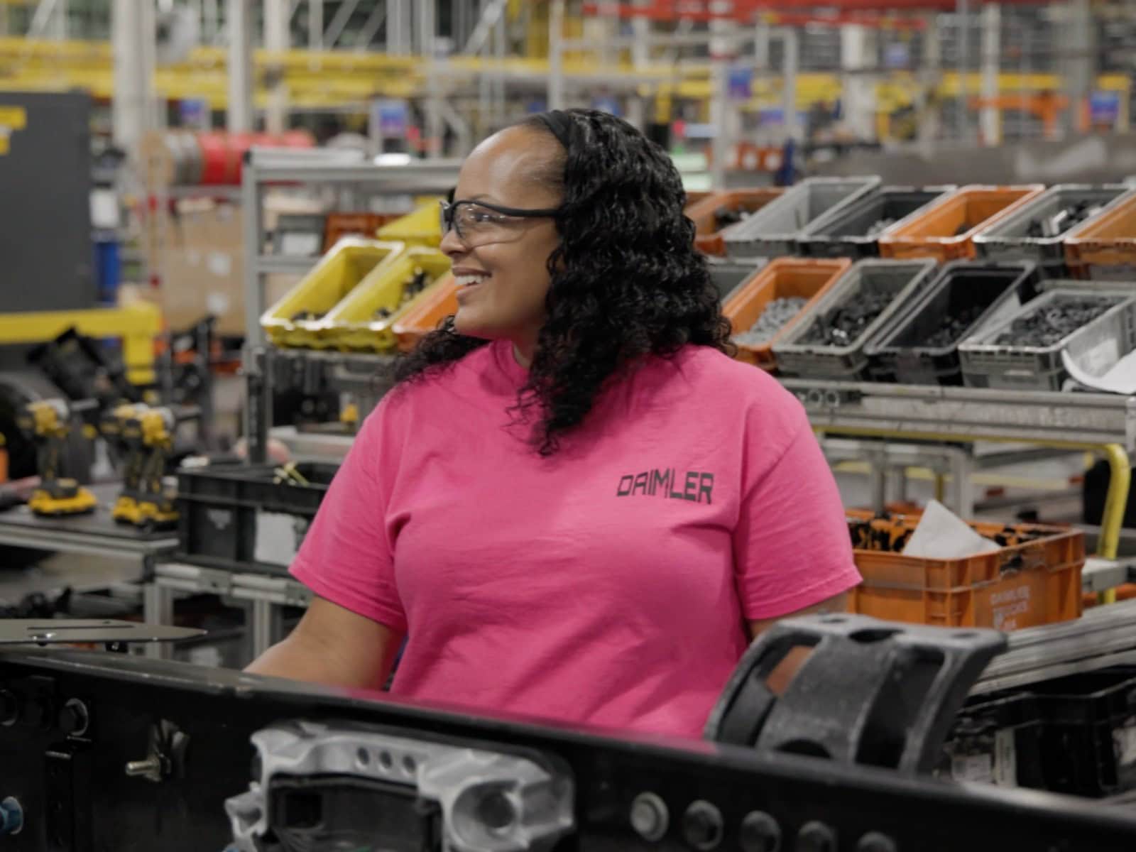 Eine Arbeiterin von Daimler Truck North America steht in der Nahaufnahme in einer Produktionshalle, umgeben von Lkw-Komponenten und Materialkisten