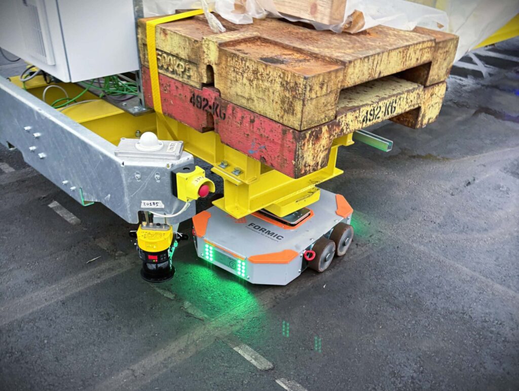 Ein Schwerlast-Schwarmfahrzeug des Karlsruher Start-ups Formic in Form eines kompakten AGV unter einer Ecke einer zu Testzwecken transportierten Ballastbrücke