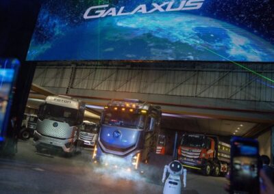 Report: China-Marke Foton auf Exportkurs Eine große, abgedunkelte Bühne mit mehreren schweren Lkw der chinesischen Marke Foton; im Vordergrund filmen Zuschauer mit ihren Mobiltelefonen