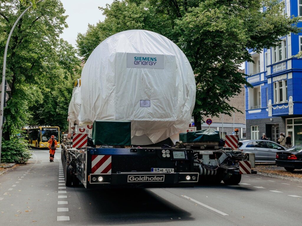 Eine rund 430 Tonnen schwere Gasturbine von Siemens Energy wird auf einem Goldhofer-Selbstfahrer mit elektrischem Powerpack durch Berlin transportiert; am Straßenrand parken Autos, im Hintergrund fährt ein Bus