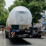 Eine rund 430 Tonnen schwere Gasturbine von Siemens Energy wird auf einem Goldhofer-Selbstfahrer mit elektrischem Powerpack durch Berlin transportiert; am Straßenrand parken Autos, im Hintergrund fährt ein Bus