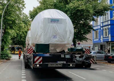 Einsatzpremiere für E-Powerpack von Goldhofer Eine rund 430 Tonnen schwere Gasturbine von Siemens Energy wird auf einem Goldhofer-Selbstfahrer mit elektrischem Powerpack durch Berlin transportiert; am Straßenrand parken Autos, im Hintergrund fährt ein Bus