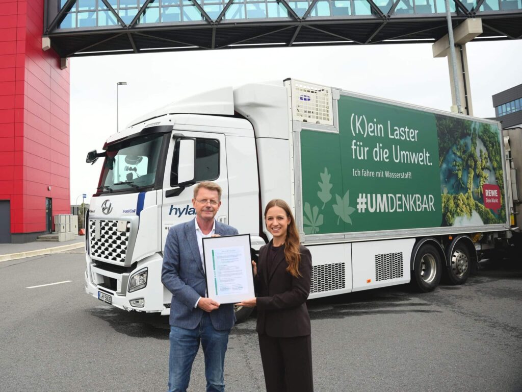 Gerd-Wilhelm Brüning (Logistikleiter REWE Nord) und Dr. Sara Schiffer (Geschäftsführerin Hylane) bei der Übergabe eines CO2-Nachweises; beide stehend vor einem Brennstoffzellen-Lkw Hyundai Xcient Fuel Cell
