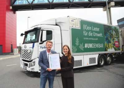 Hylane: Nachgewiesene CO2-Ersparnis mit Wasserstoff Gerd-Wilhelm Brüning (Logistikleiter REWE Nord) und Dr. Sara Schiffer (Geschäftsführerin Hylane) bei der Übergabe eines CO2-Nachweises; beide stehend vor einem Brennstoffzellen-Lkw Hyundai Xcient Fuel Cell