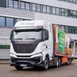 Sattelzug mit Sattelauflieger des Pharma-Unternehmens Teva und einer Brennstoffzellen-Sattelzugmaschine Iveco S‑eWay Fuel Cell aus der Mietflotte von Hylane; Fahrzeug parkt vor einem Logistikgebäude