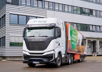Sattelzug mit Sattelauflieger des Pharma-Unternehmens Teva und einer Brennstoffzellen-Sattelzugmaschine Iveco S‑eWay Fuel Cell aus der Mietflotte von Hylane; Fahrzeug parkt vor einem Logistikgebäude