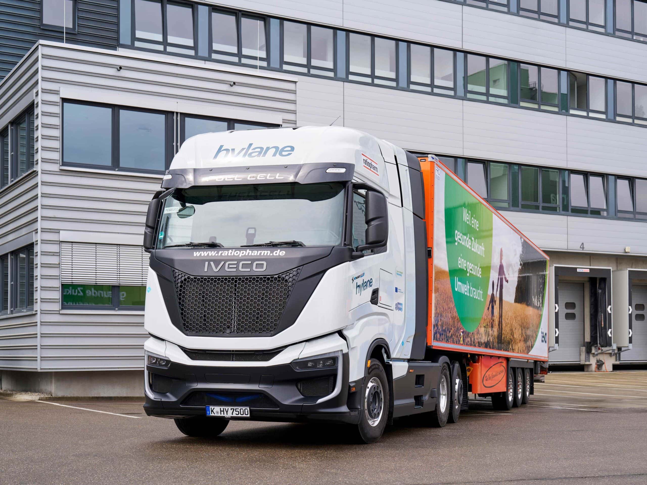 Sattelzug mit Sattelauflieger des Pharma-Unternehmens Teva und einer Brennstoffzellen-Sattelzugmaschine Iveco S‑eWay Fuel Cell aus der Mietflotte von Hylane; Fahrzeug parkt vor einem Logistikgebäude