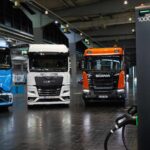 Elektro-Lkw von Mercedes-Benz Trucks, MAN und Scania nebeneinander in einer Halle, im Vordergrund rechts eine Megawatt-Ladesäule von Milence
