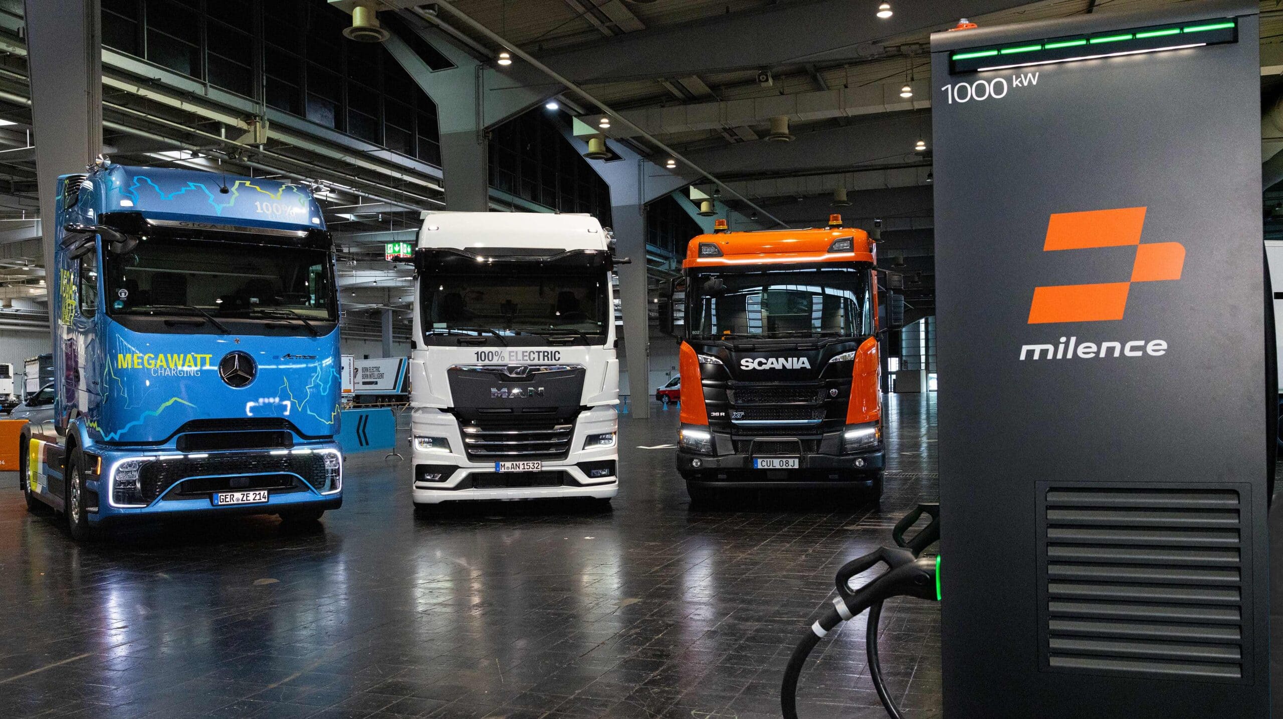 Elektro-Lkw von Mercedes-Benz Trucks, MAN und Scania nebeneinander in einer Halle, im Vordergrund rechts eine Megawatt-Ladesäule von Milence