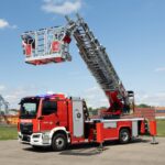 Drehleiterfahrzeug für die Feuerwehr auf einer Stellfläche, Stützen und Rettungskorb sind ausgefahren; es ist ein vollelektrisches Fahrzeug mit Aufbau von Rosenbauer auf einem MAN eTGM