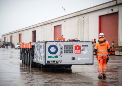 Emissionsfreier Schwertransport mit Scheuerle E-SPMT Selbstfahrender Scheuerle-Modultransporter mit elektrischer PowerPack Unit (ePPU), begleitet von einem Arbeiter im Hafeneinsatz