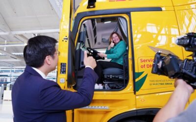 Antje Huber (DHL Freight) und Chao Liu (Gründer & CEO Superpanther) bei der feierlichen Fahrzeugübergabe eines E-Lkw eTopas 600 in Steyr; Antje Huber sitzt bei offener Tür im Fahrerhaus des Lkw, Chao Liu steht mit Daumen-hoch-Geste davor