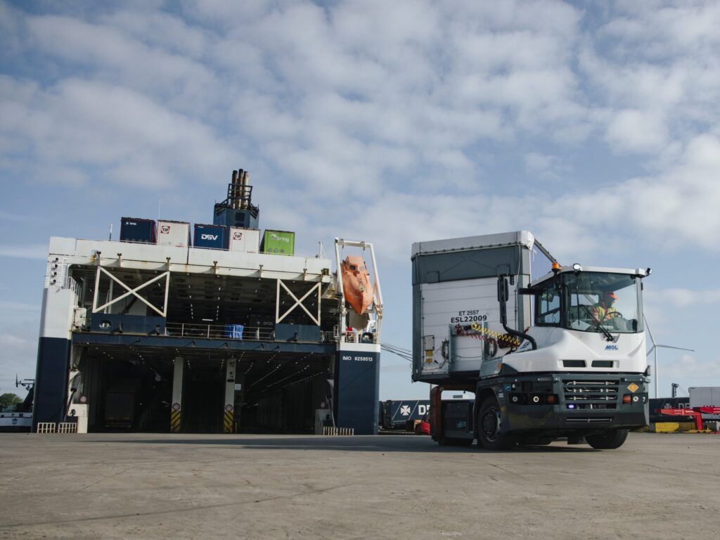 Batterieelektrisch angetriebener RoRo-Traktor von MOL in einem Hafen, im Hintergrund ein angelandetes Fährschiff